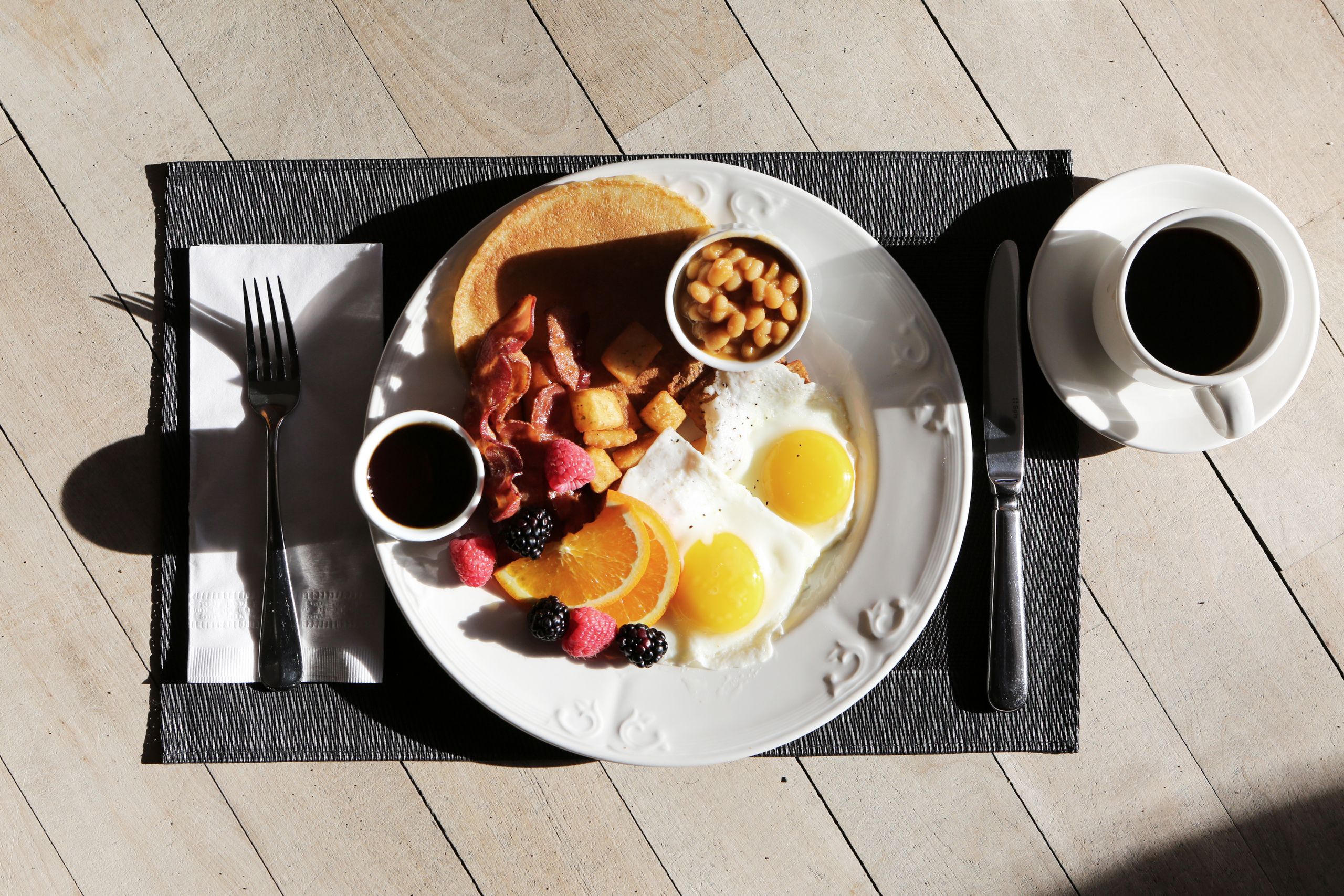 breakfast spread featuring eggs, bacon, fruit, and coffee on a sunlit table.